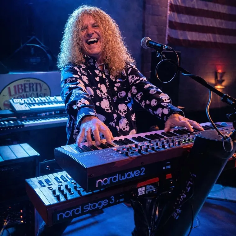 Chip McGuer laughing over a Nord Stage 3 and Nord Wave 2, an American flag on the wall behind him